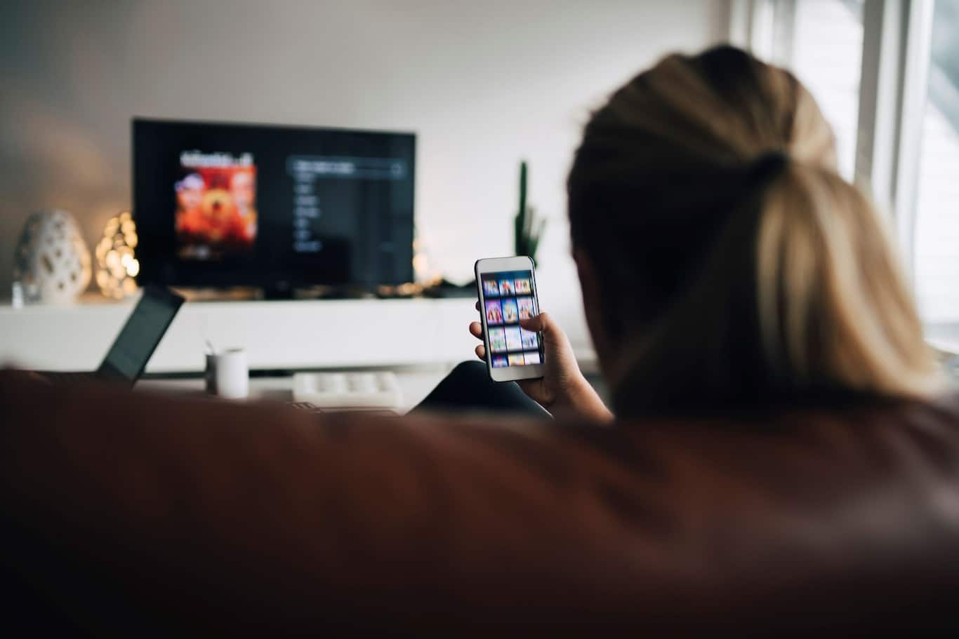 mladá žena sedí pred televíznom obrazovkou a sleduje smartfón
