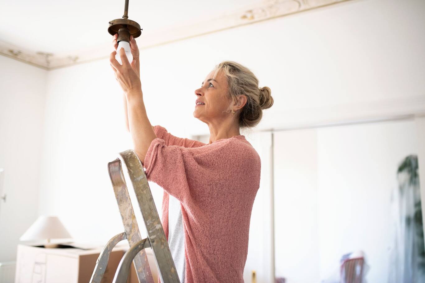 Žena, ktorá stojí na rebríku a vymieňa bežnú žiarovku za LED žiarovku