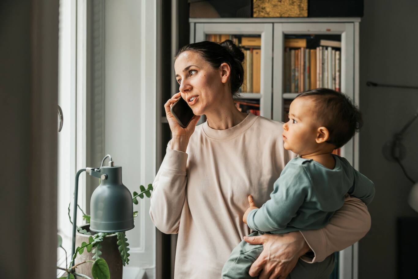 Osoba drží dieťa a telefonuje pri okne doma. Zisťuje informácie o svojom dôchodkovom sporení v 2. pilieri.